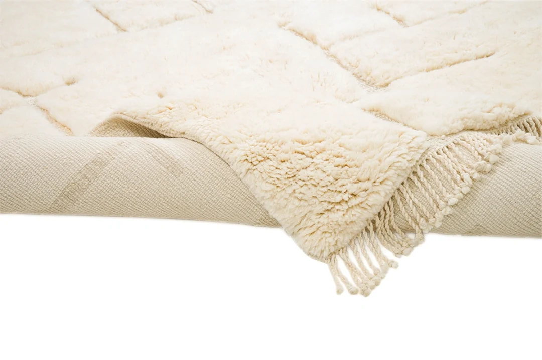A cream-colored textured rug with a fringe on one end is partially folded to show both the plush pile and woven underside.