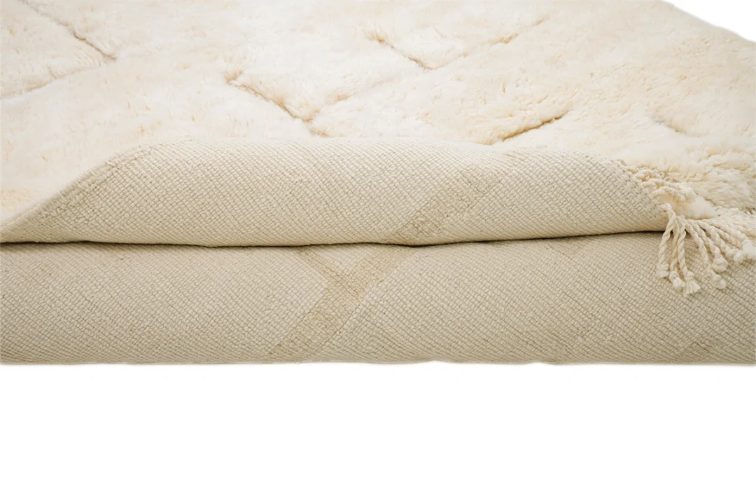 A close-up view of a folded cream-colored textured rug with a plush, fluffy surface and tassels on one corner.