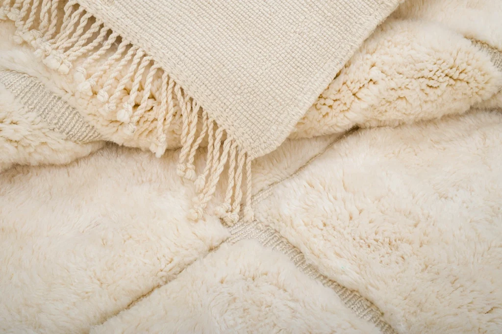 A close-up of a cream-colored woven throw rug with fringed edges lying on top of a soft, fluffy, light beige comforter.