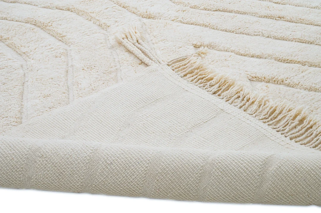 A close-up of a cream-colored textured rug with a corner flipped over, showing the underside and fringe detail.