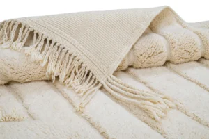 A cream-colored, textured blanket with fringed edges is folded neatly, showing both its woven and plush sides.