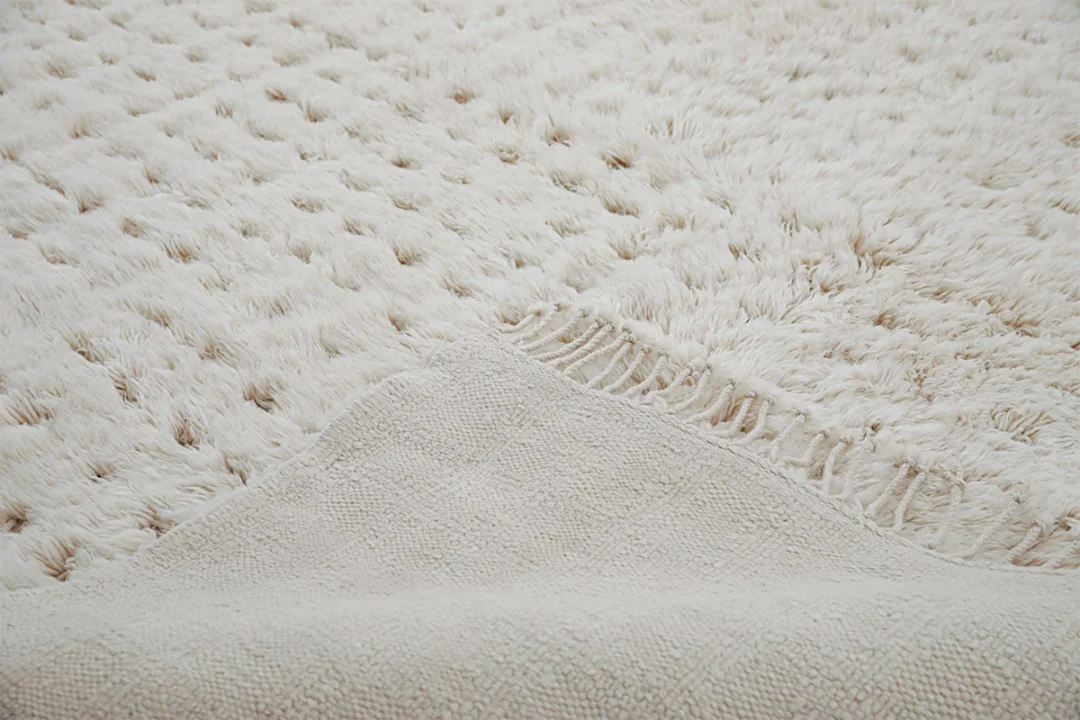 Close-up of a textured, off-white rug with a corner lifted to show the rug backing and different pile patterns.