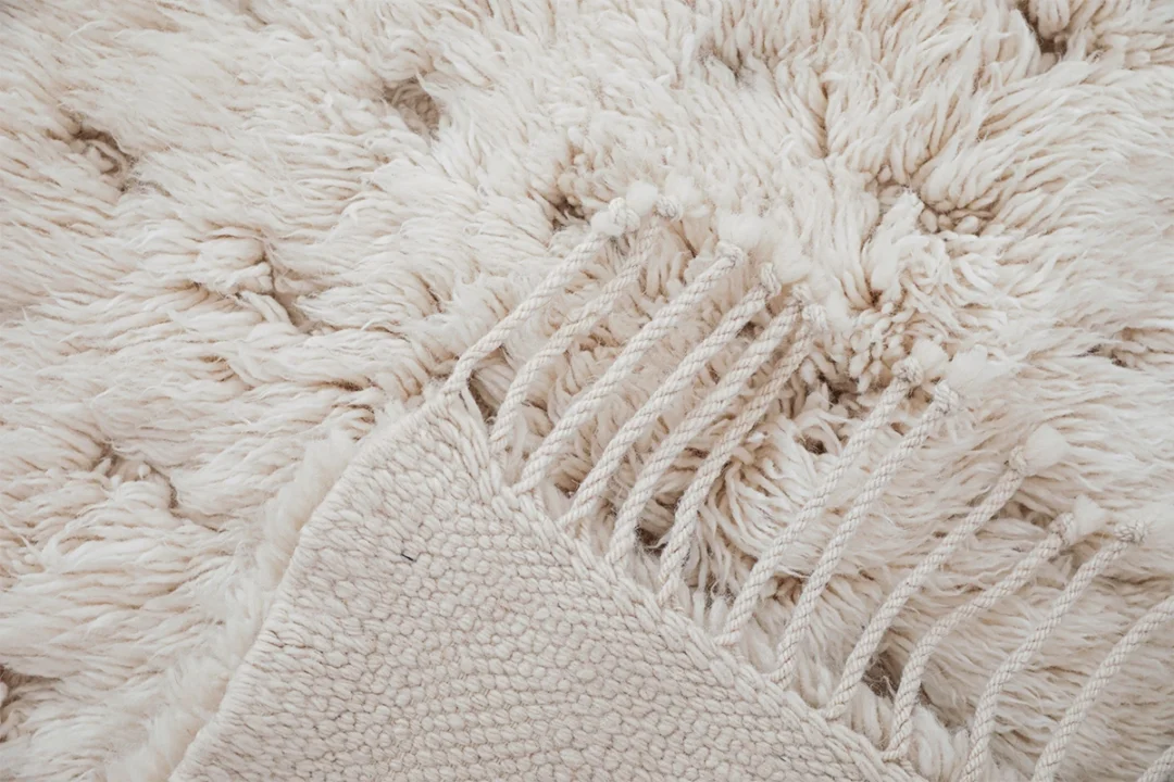 Close-up of a cream-colored shaggy rug with visible fringe and a folded corner showing the rug’s underside.