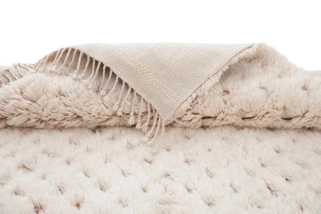 A beige woven throw rug with fringed edges is laid over a thick, plush, cream-colored textured rug.