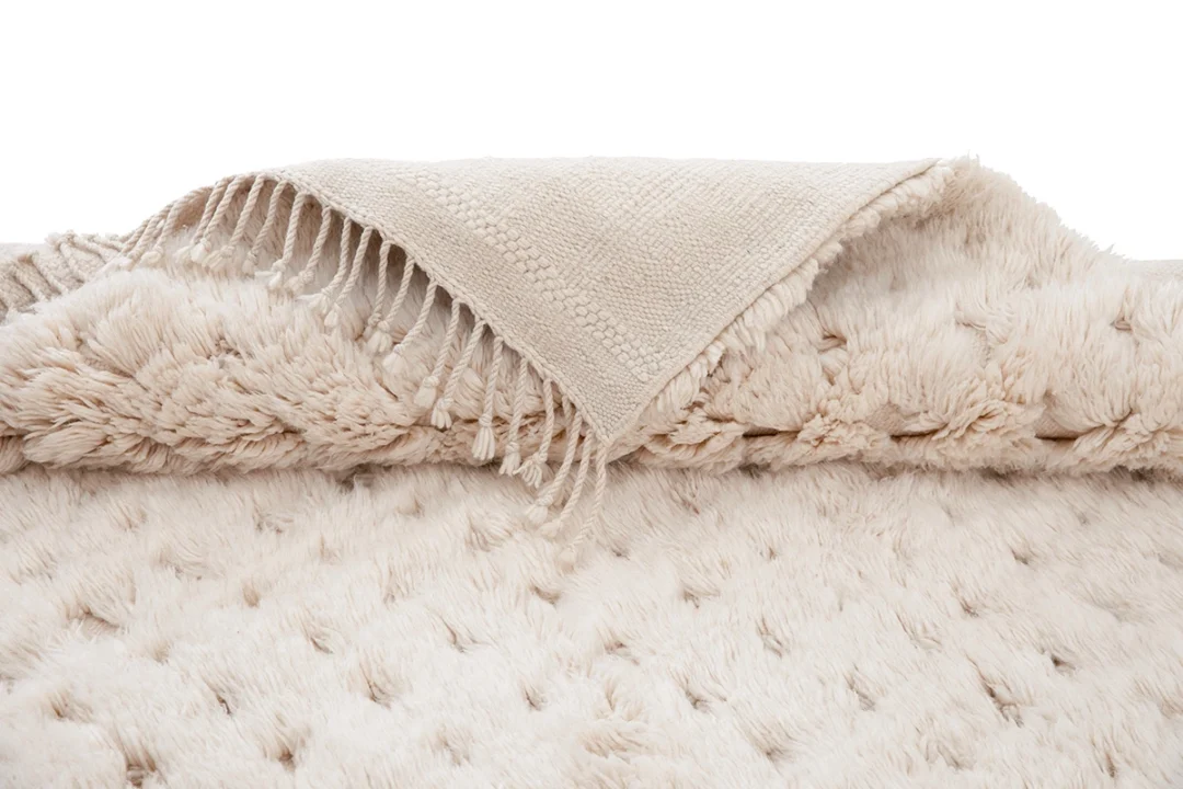 A beige woven throw rug with fringed edges is laid over a thick, plush, cream-colored textured rug.