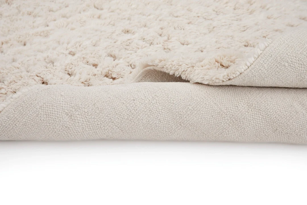 Close-up of a light beige, textured, fluffy rug with one corner folded over, revealing the woven underside.