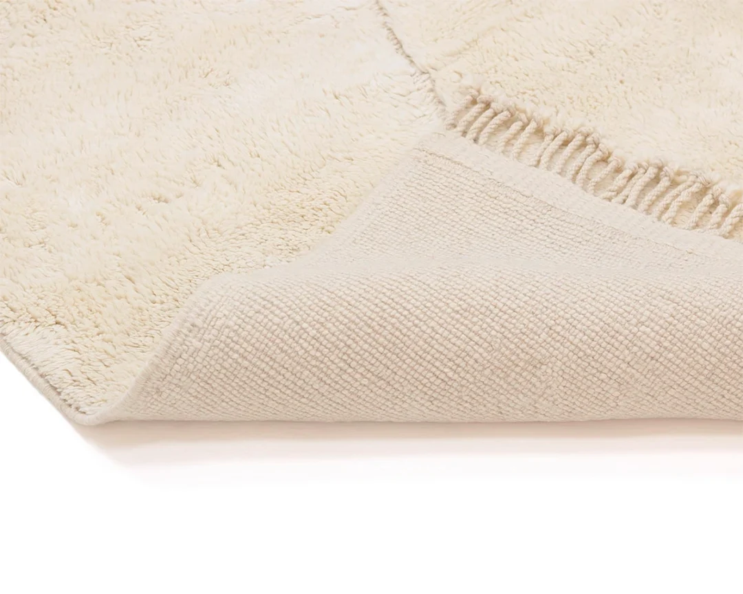 A close-up of a cream-colored textured rug with one corner folded over, showing the woven underside and soft surface.
