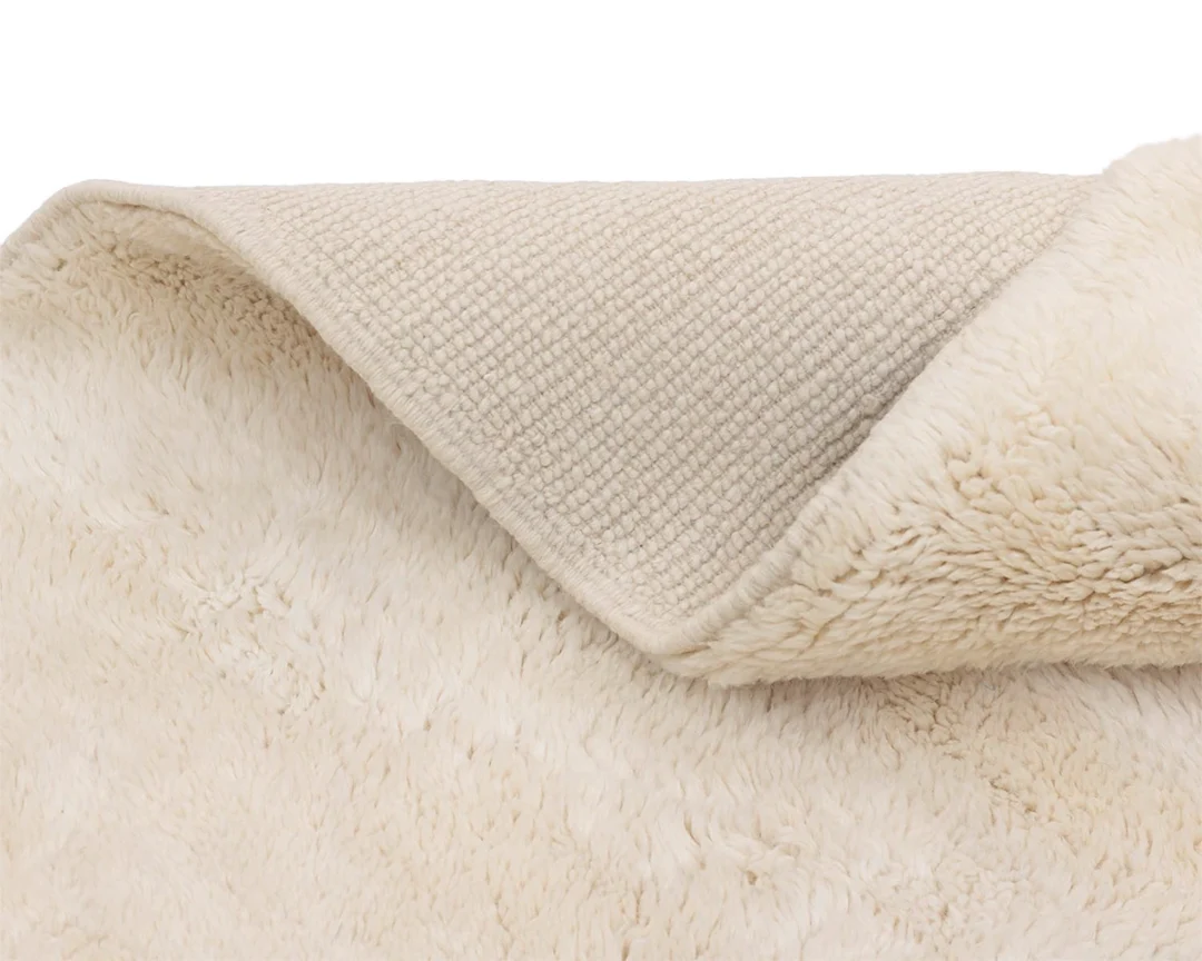 A close-up view of a cream-colored, fluffy rug with the corner folded over, revealing its textured woven underside.