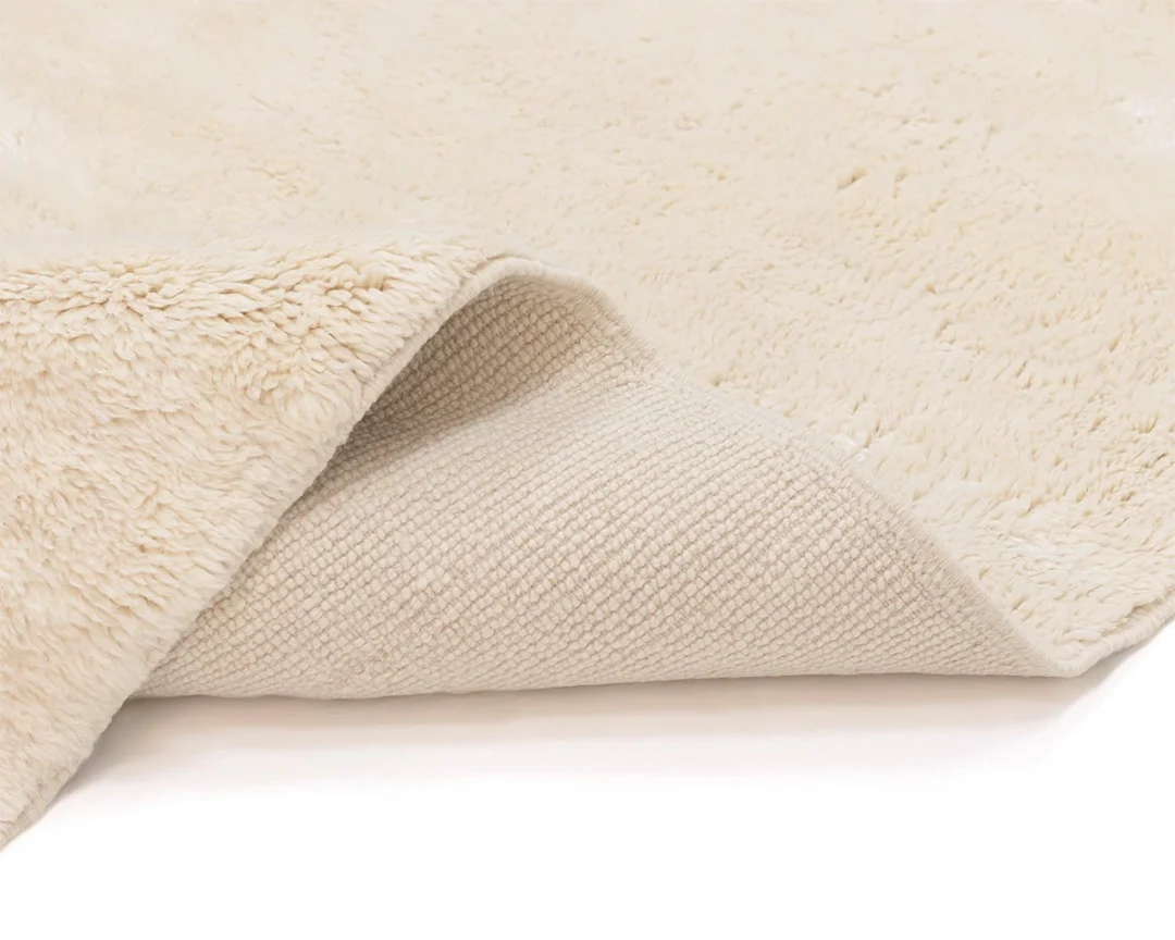 A close-up view of a cream-colored, fluffy rug with one corner folded over, showing the textured backing underneath.