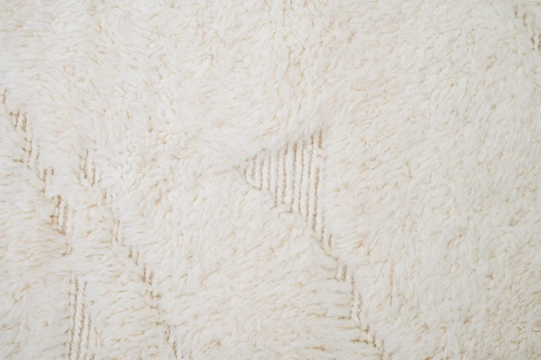 Close-up of a cream-colored, textured, fluffy rug with subtle geometric patterns and a small area showing the underlying weave.