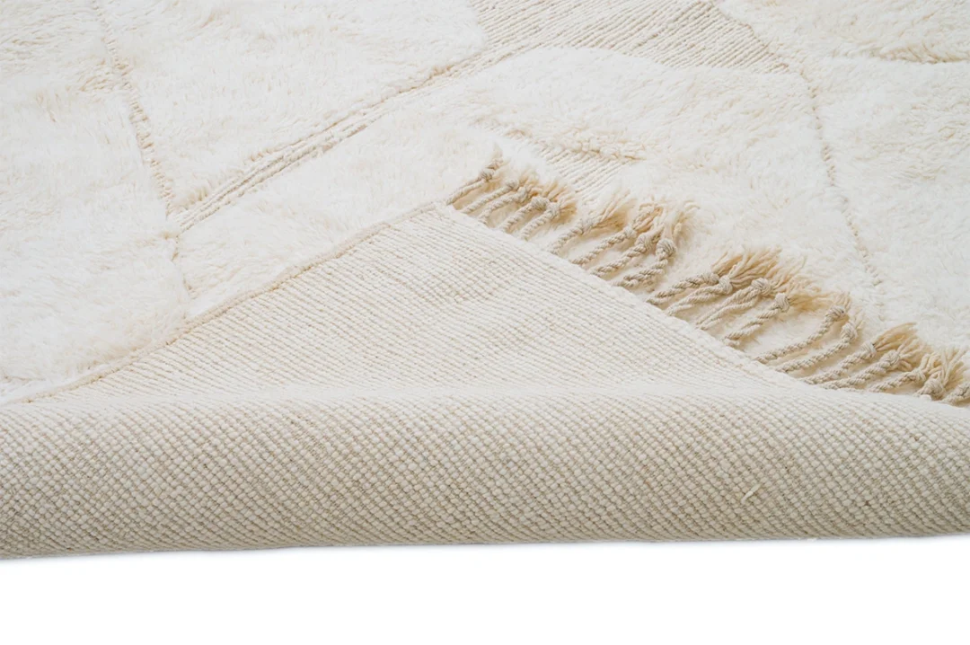 Close-up of a cream-colored textured rug with a visible corner flipped to show the woven underside and fringe detailing.