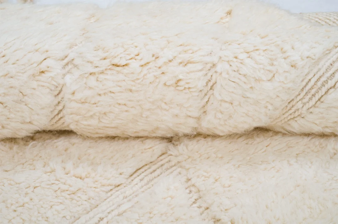 Close-up view of a folded, off-white, fluffy textured fabric or rug with visible stitching lines.