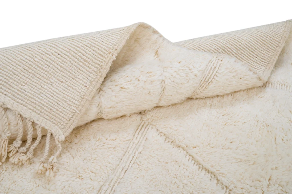 A close-up of a cream-colored, textured area rug with decorative fringe and a folded corner showing the woven underside.