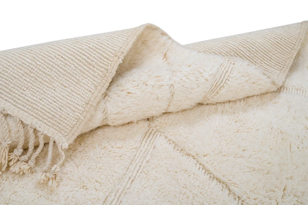 A close-up of a cream-colored, textured area rug with decorative fringe and a folded corner showing the woven underside.