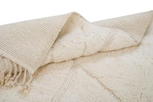 A close-up of a cream-colored, textured area rug with decorative fringe and a folded corner showing the woven underside.
