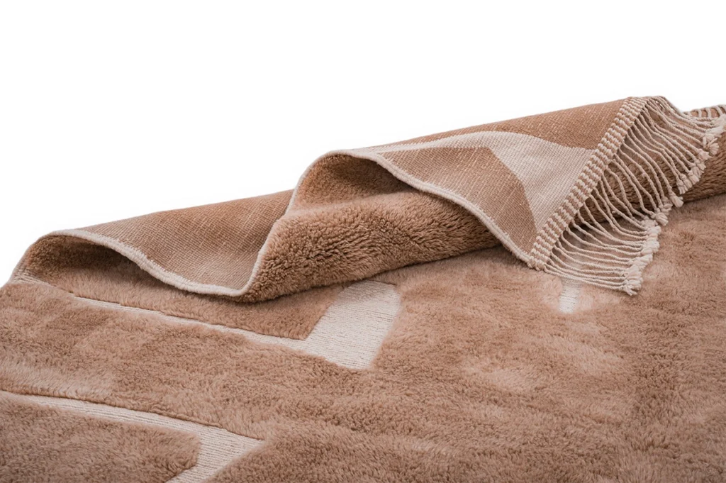 A close-up of a folded, light brown and beige blanket with a geometric pattern and fringed edges on a white background.