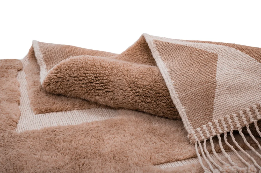 A close-up of a soft, beige and cream textured throw blanket with fringe on one edge, partially folded to show both the fluffy and woven sides.