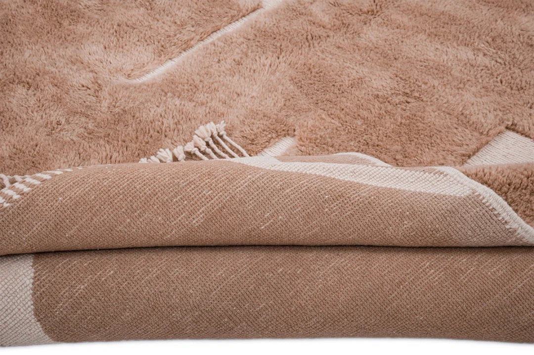 A close-up view of a folded beige textured rug or blanket with a soft, plush surface and visible fringes.