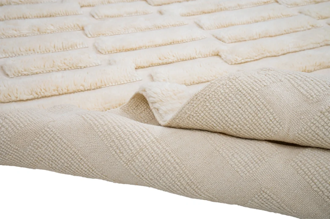 A close-up of a textured cream-colored rug with raised rectangular patterns and a folded corner showing the underside.