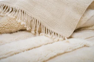 A close-up of a textured beige woven blanket with tassels partially covering a fluffy, cream-colored faux fur surface.