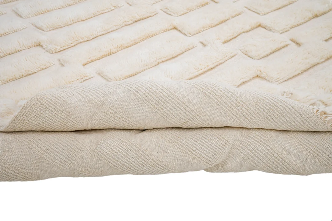 A close-up of a folded beige textured rug with a geometric pattern, showing both the front and the edge.