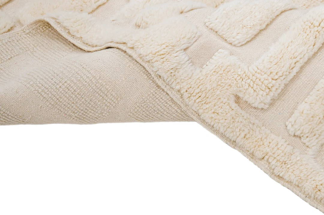 Close-up of a beige textured rug with raised geometric patterns and a soft, plush surface, shown partially folded over on a white background.