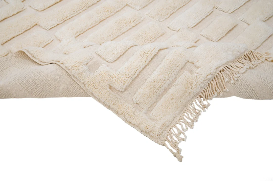 A beige area rug with a raised geometric pattern and fringed edges, partially folded over to show texture and thickness.