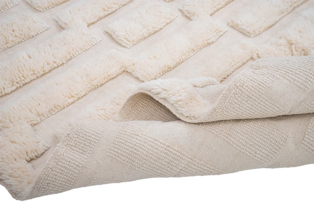 A close-up of a cream-colored textured rug with raised rectangular patterns and a folded corner showing the underside.
