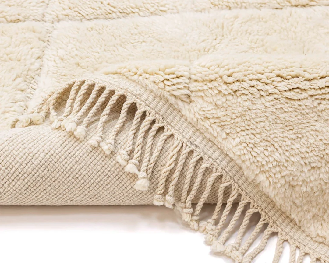 A close-up of a cream-colored textured rug with a fringe edge, partially folded to show both the plush surface and woven underside.