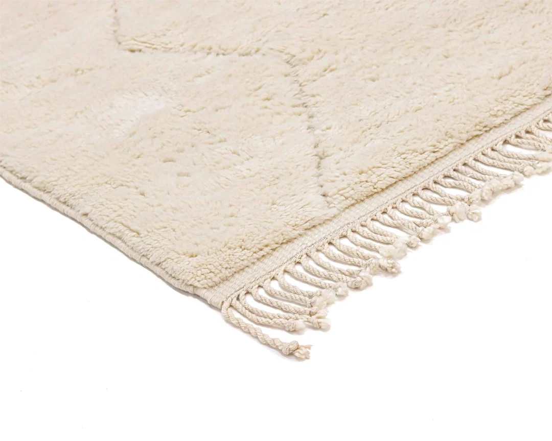 A close-up of a beige, textured rug with a subtle geometric pattern and fringed tassels along the edge, shown on a white background.