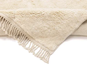 A close-up of a cream-colored, textured throw blanket with fringed tassels along the edge, folded over on a white background.