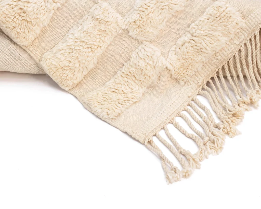 A beige textured throw blanket with raised patterns and fringe detail along the edge is shown partially folded on a white background.