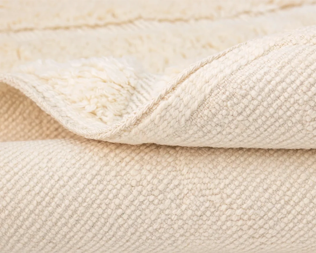 Close-up view of a folded, textured cream-colored knotted rug with visible fibers and subtle patterns.