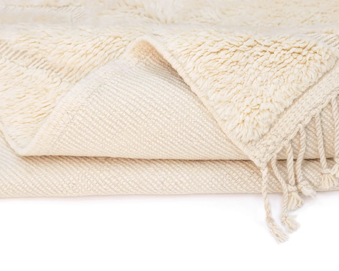 A folded cream-colored textured rug with tassels on the edge, showing both a soft, fluffy side and a woven side.