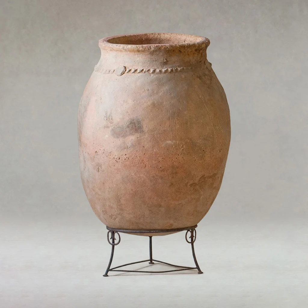 Large, weathered clay jar with a rounded body and narrow neck, featuring a simple raised band, displayed on a metal stand against a plain background.