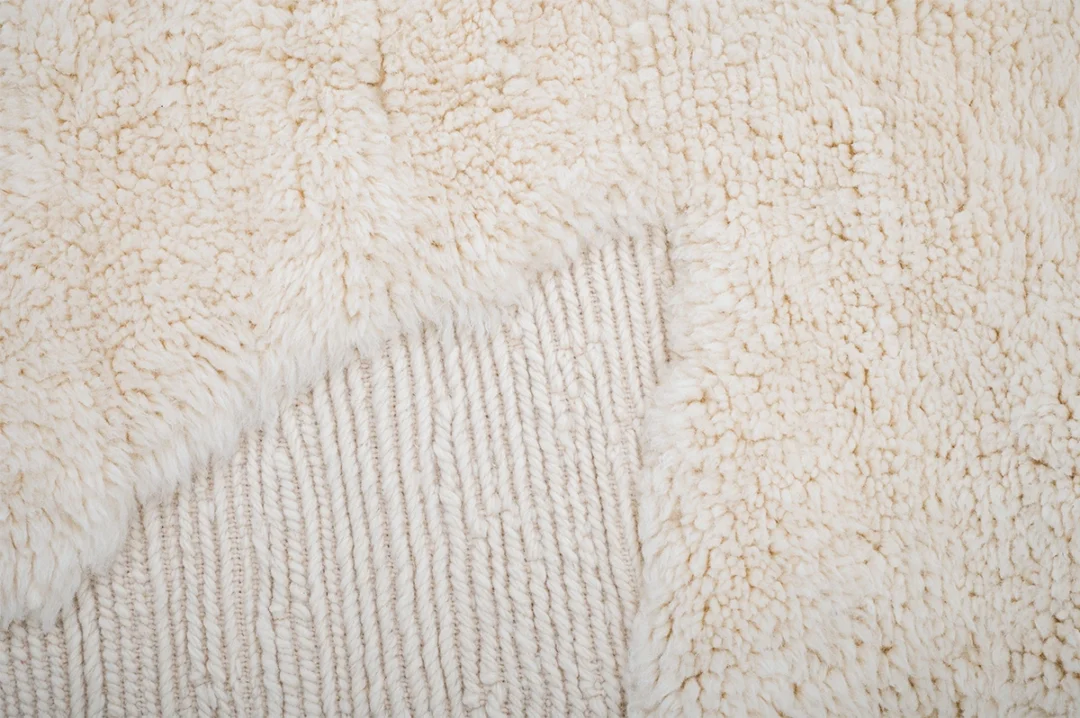 Close-up of a cream-colored textured rug with a soft, fluffy surface partially pulled back to reveal a ribbed knit pattern underneath.