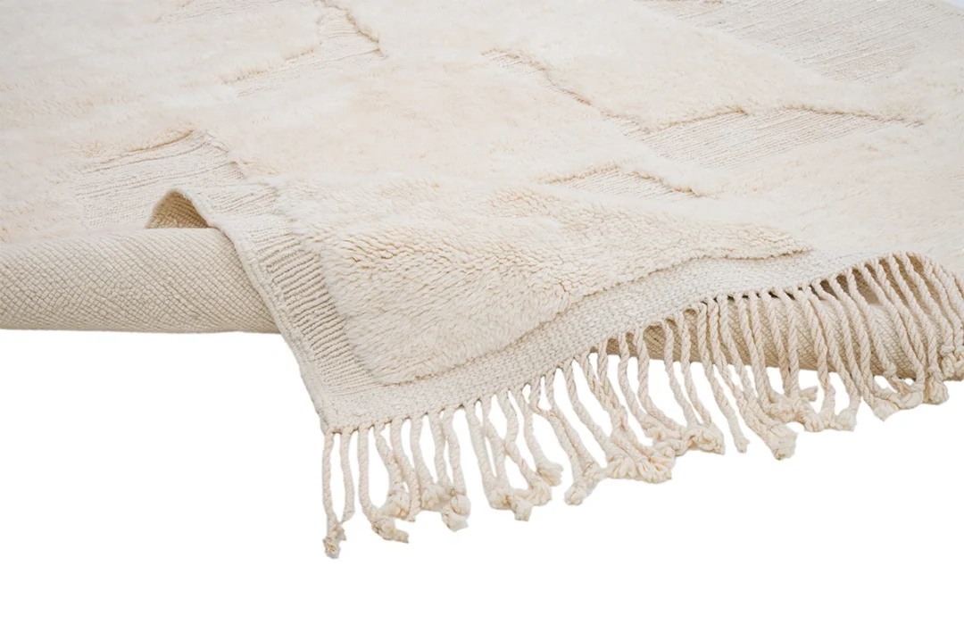 A cream-colored woven rug with textured patterns and fringed edges, partially rolled and laid on a white surface.
