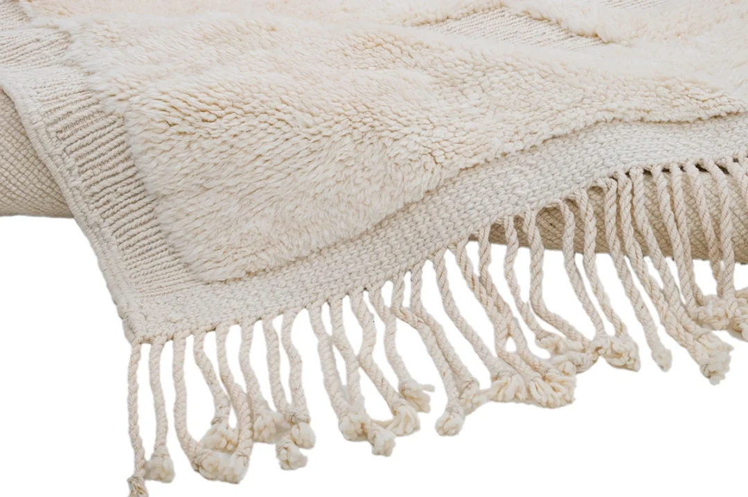 A close-up view of a cream-colored woven rug with textured patterns and twisted fringe along the edge.