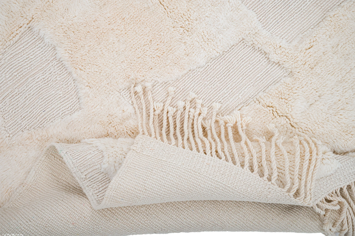 Close-up of a beige textured rug with a geometric pattern, fringed edges, and one corner folded to show the woven underside.