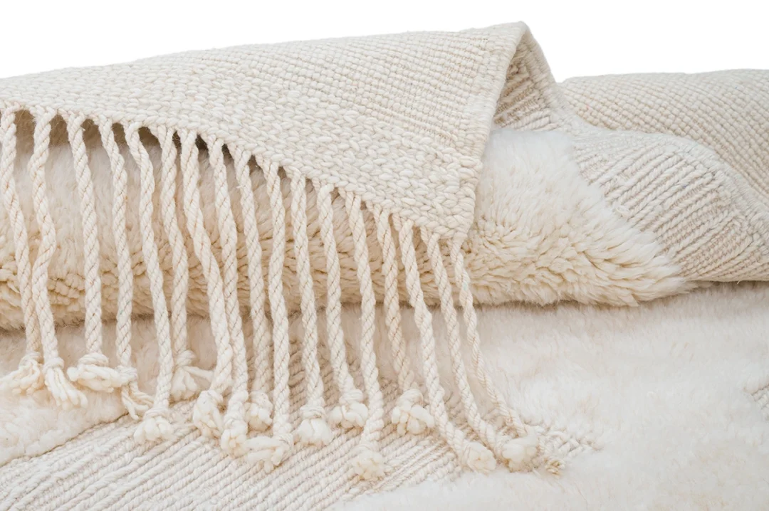A folded cream-colored woven throw rug with thick fringe tassels lies on top of a fluffy, textured white surface.