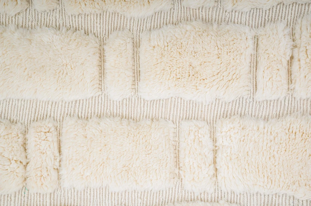 A close-up view of a textured cream-colored rug with rectangular fluffy sections and a woven background.