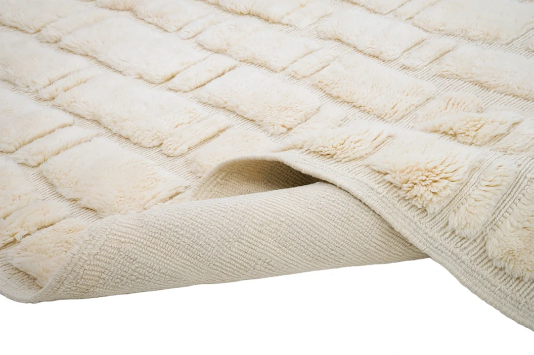 A close-up of a cream-colored textured rug with a raised rectangular pattern, showing the corner slightly lifted to reveal the underside.