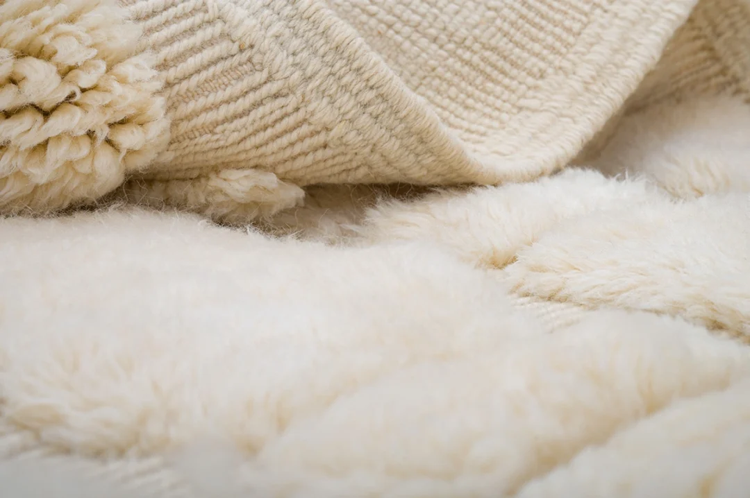Close-up of a cream-colored, fluffy wool rug with a textured woven edge partially folded over a soft surface.
