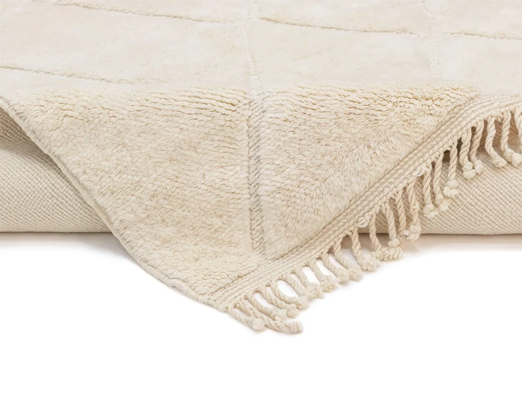 A close-up of a cream-colored, textured throw blanket with fringe tassels along the edges, folded on a surface.