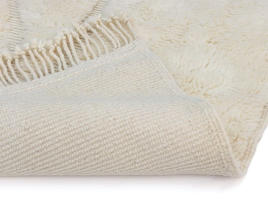 Close-up of a cream-colored textured rug with fringe edging, showing the corner flipped over to reveal the woven underside.
