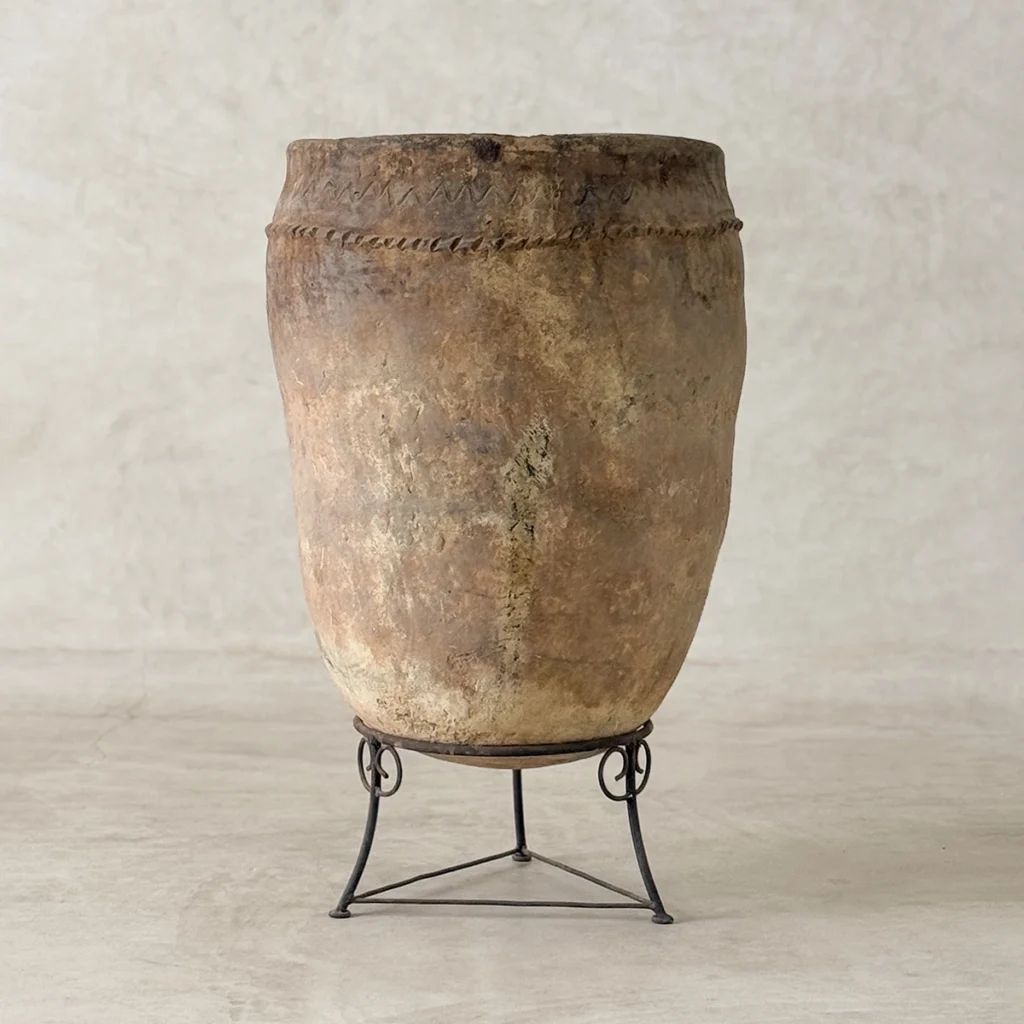 A large, weathered clay pot with a narrow mouth sits on a simple black metal stand against a plain, light-colored background.