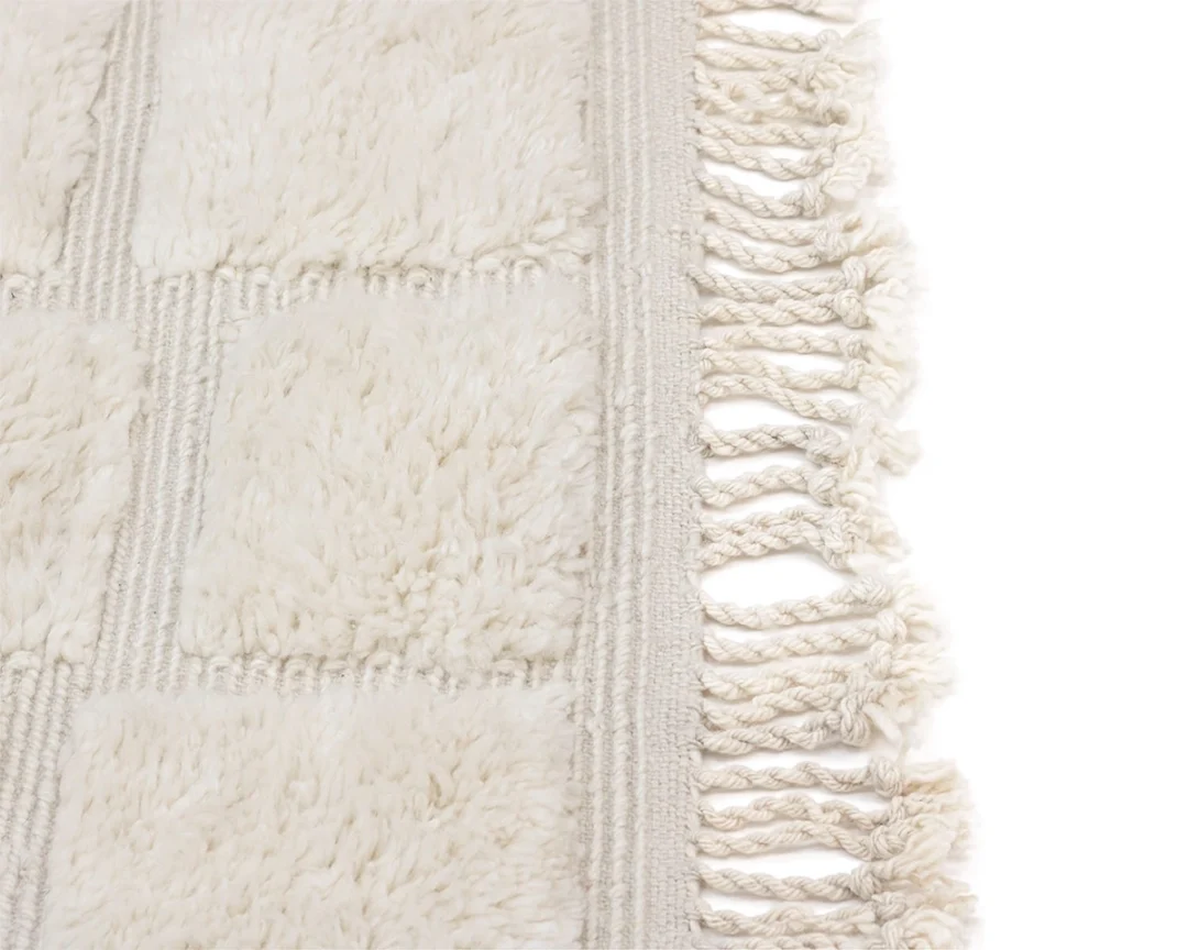 Close-up of a cream-colored textured rug with square patterns and fringed tassels along the edge, shown against a white background.