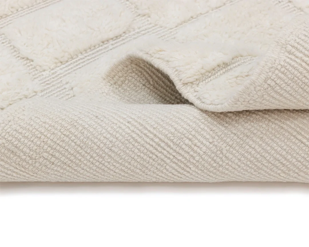 A close-up of a light beige, textured wool rug with a slightly lifted corner showing its thickness and woven underside.