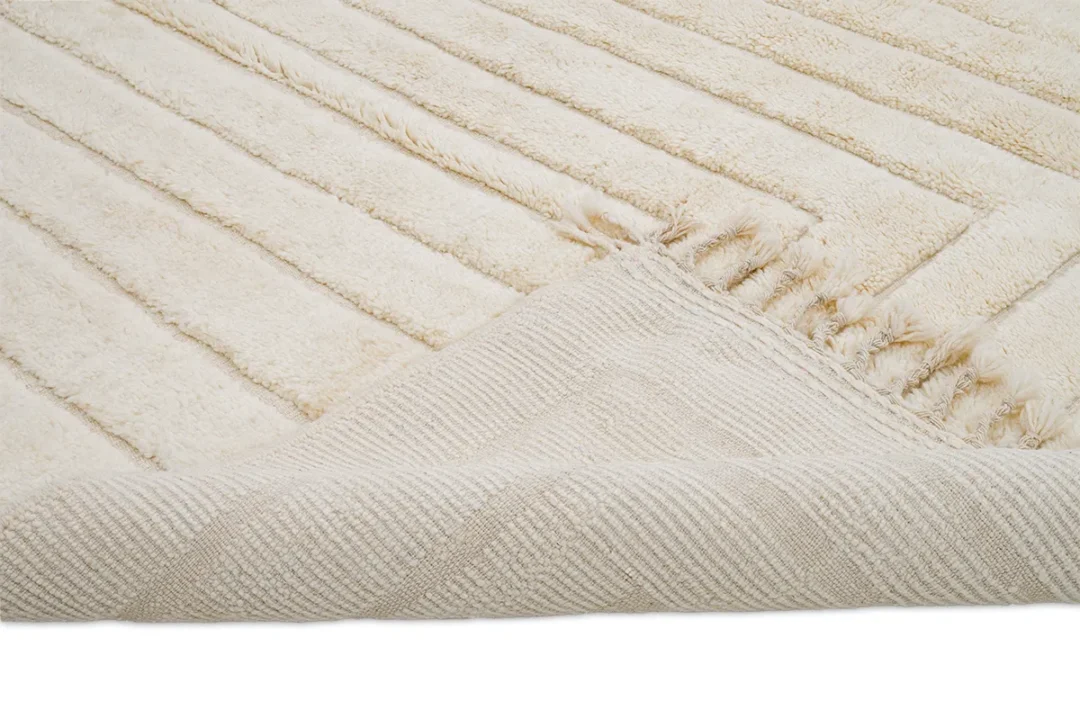 A close-up of a cream-colored textured rug with a corner folded over, showing the back side and linear pattern on the surface.