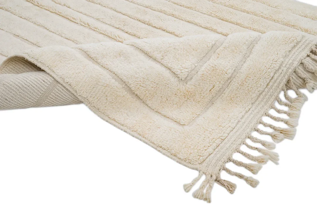 A beige textured rug with horizontal stripe patterns and tasseled fringe edges is partially folded to show both the front and back sides.